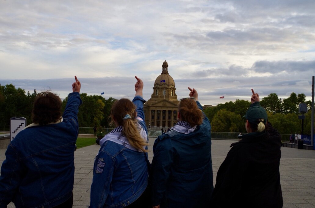 Panoramic photo of Alberta Legislature. In front of it, four people, facing the legislature, point their middle fingers towards it.