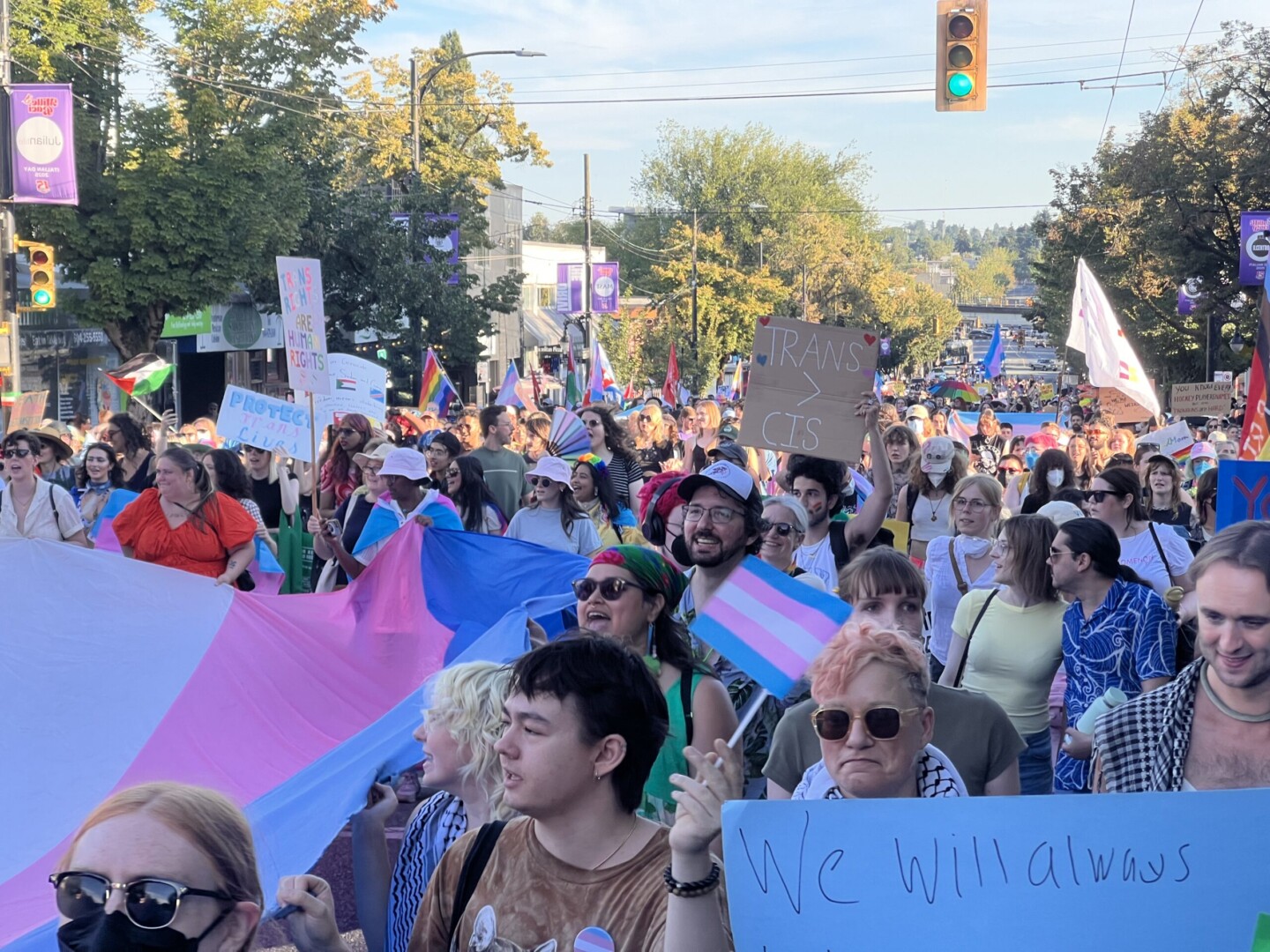 Vancouver Trans March, 1 August 2025. Giant trans flag and multiple marchers with a variety of different signs, including one in the foreground saying 