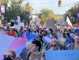 Vancouver Trans March, 1 August 2025. Giant trans flag and multiple marchers with a variety of different signs, including one in the foreground saying 